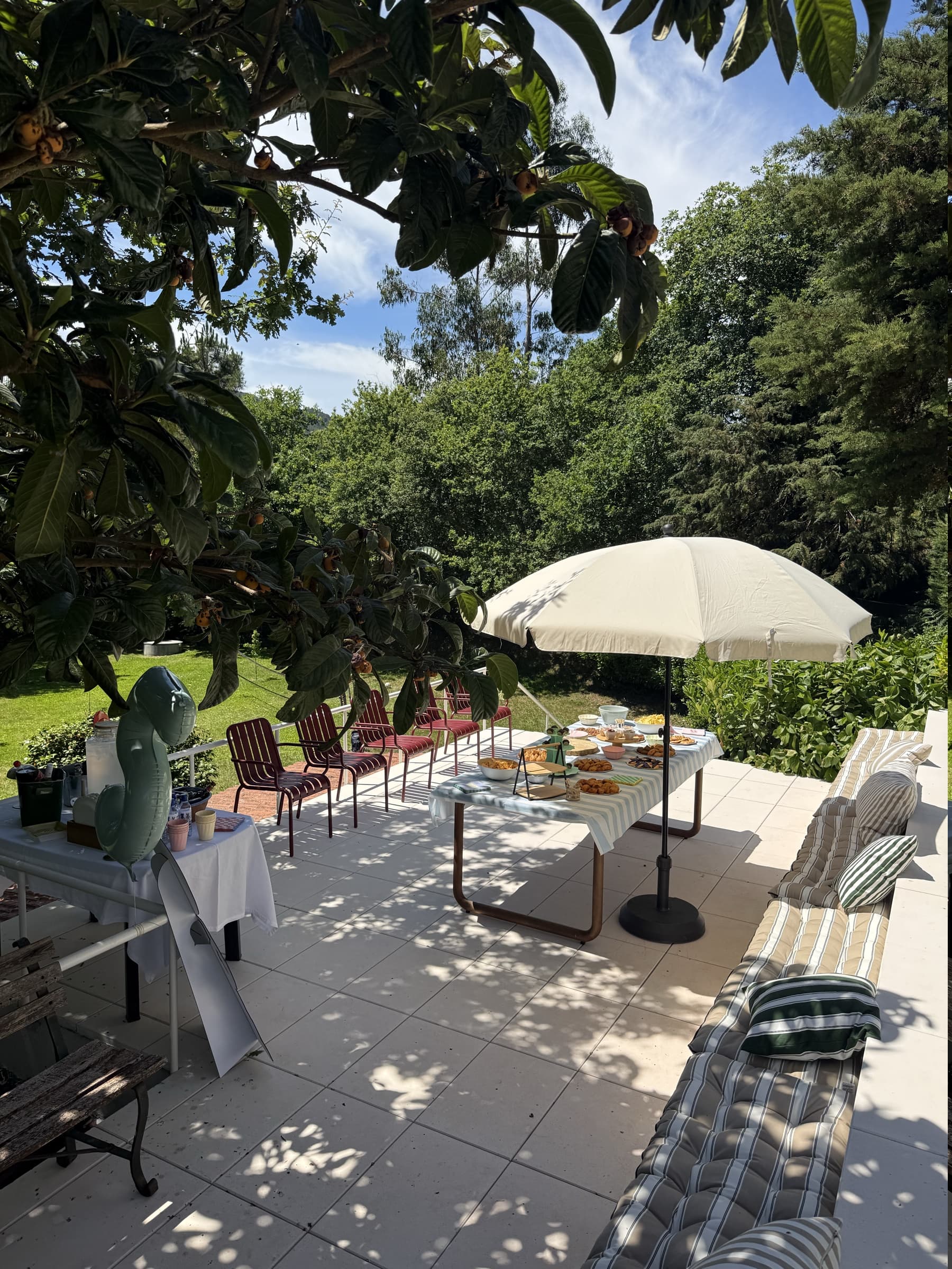 O pátio com a mesa posta e guarda-sol, debaixo dos ramos da nespereira, com espreguiçadeiras e o relvado em fundo.