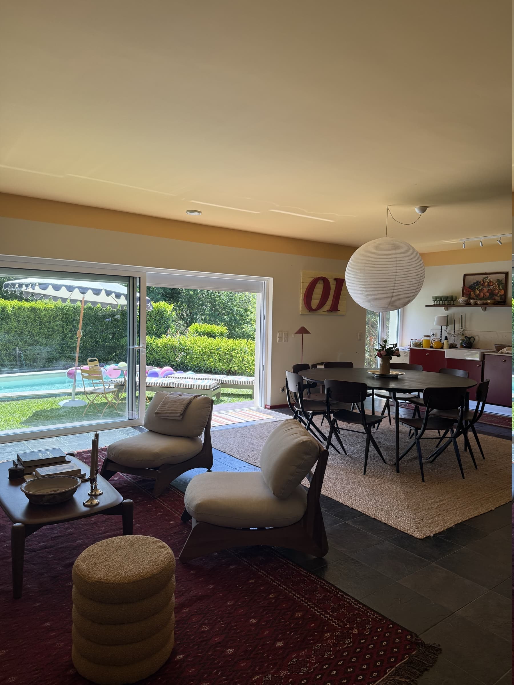 A sala em espaço aberto: zona de estar, mesa de jantar e a cozinha vermelha ao fundo, com a piscina à vista pelas portas de correr.